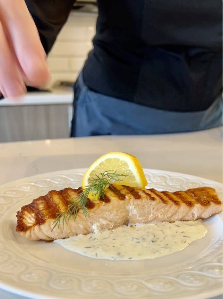 Grilled salmon fillet with lemon wedge and creamy sauce on a white plate.